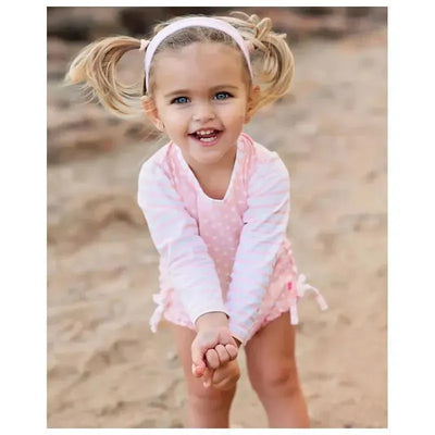 Baby girl in a pink polka dot long sleeve rash guard, smiling and playing in the sand.