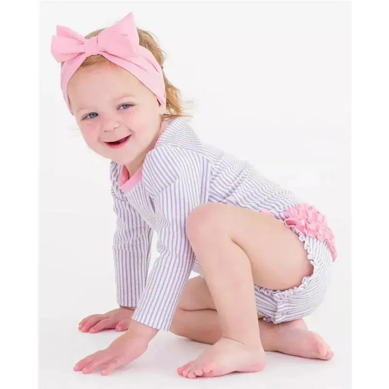 Baby girl wearing a lavender seersucker long sleeve one-piece rash guard with pink accents, smiling while kneeling.