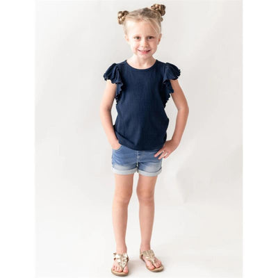 Baby girl wearing light wash stretch denim shorts and a navy top with ruffle sleeves, smiling and standing with hands on hips.