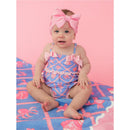 Baby girl wearing a pink and blue tropical one-piece swimsuit with bows and ruffles, sitting on a colorful beach towel.