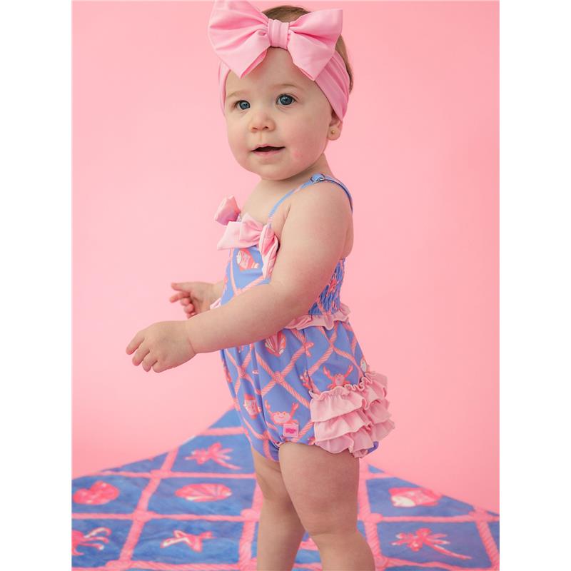 Baby girl wearing a tropical-patterned one-piece swimsuit with bows and ruffles, set against a pink background.