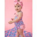 Baby girl wearing a tropical-patterned one-piece swimsuit with bows and ruffles, set against a pink background.