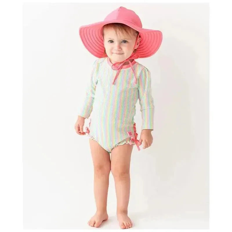 Baby girl wearing a colorful striped long-sleeve swimsuit with a pink ruffle swim hat and smiling.