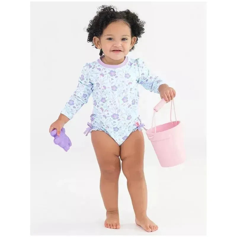 Toddler wearing a floral long sleeve rash guard, holding a pink bucket and a purple toy, smiling at the camera.