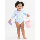Toddler wearing a floral long sleeve rash guard, holding a pink bucket and a purple toy, smiling at the camera.