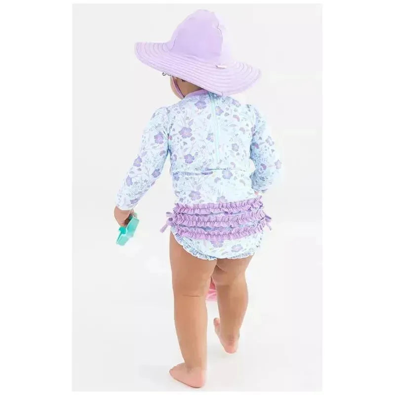 Toddler wearing a floral long sleeve rash guard and ruffled swim bottoms, holding a water toy and wearing a purple sun hat.