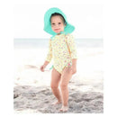Toddler wearing a pastel yellow swimsuit with colorful patterns and an island blue sun hat, standing on a sandy beach.