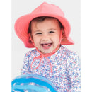 Kids swim hat by Rufflebutts in bubblegum pink worn by a smiling child holding a blue inflatable beach ball.