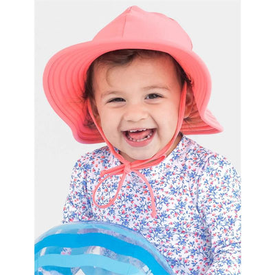 Kids swim hat by Rufflebutts in bubblegum pink worn by a smiling child holding a blue inflatable beach ball.