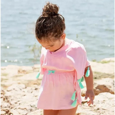 Pink tassel kaftan cover-up by Rufflebutts worn by a young girl, standing by a lake.
