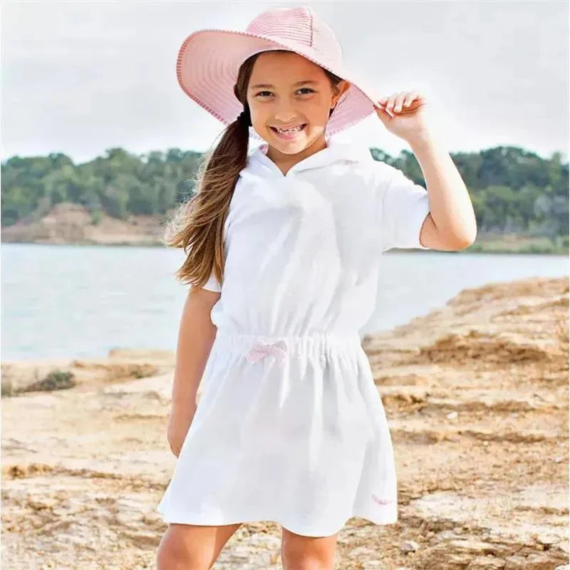 Child wearing a white and pink seersucker terry cover-up with a hood, smiling by the water in a sunhat.