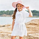 Child wearing a white and pink seersucker terry cover-up with a hood, smiling by the water in a sunhat.