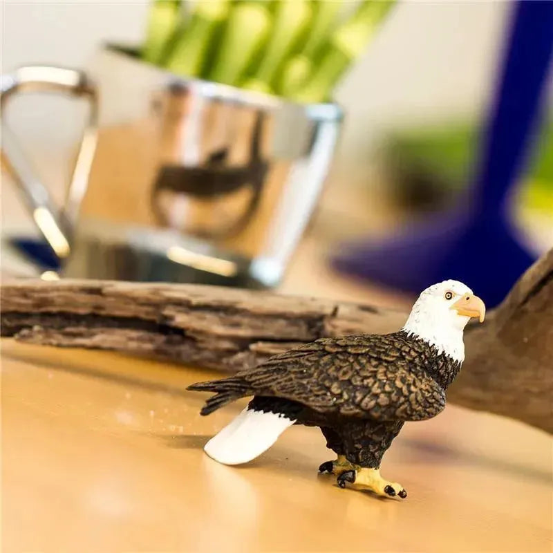 Bald Eagle figurine by Safari Ltd., detailed with brown feathers and a white head, positioned on a wooden surface.
