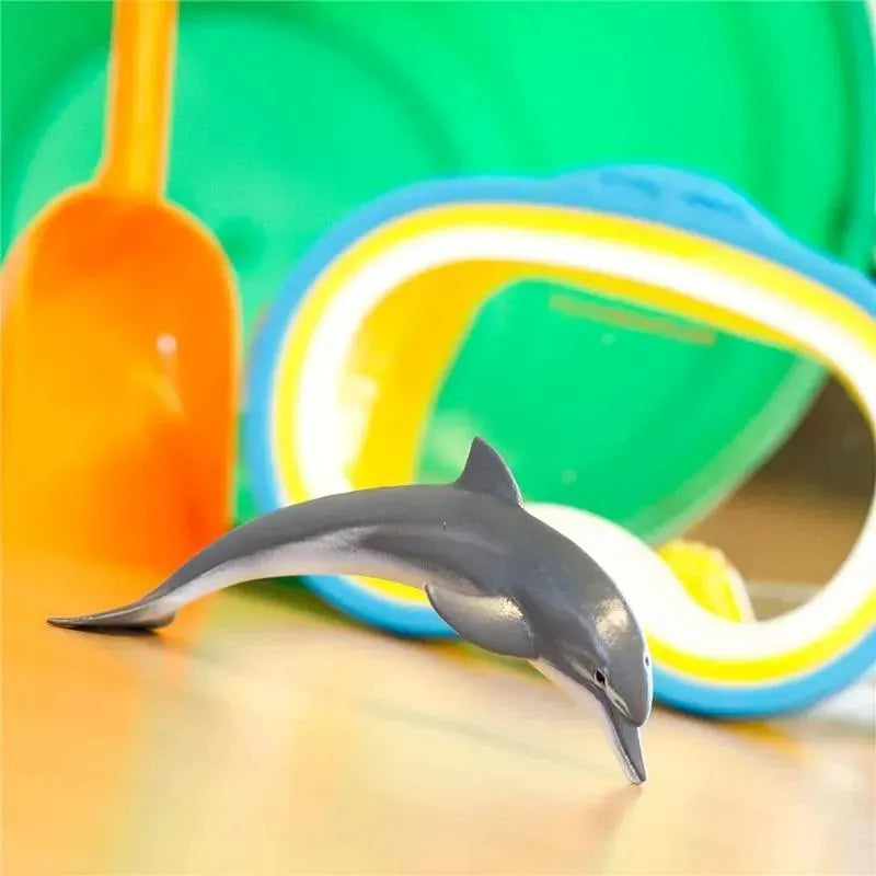 Lifelike 4.5-inch dolphin figurine positioned in front of colorful beach toys including a shovel and goggles.