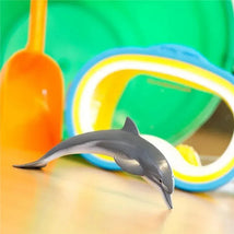 Lifelike 4.5-inch dolphin figurine positioned in front of colorful beach toys including a shovel and goggles.