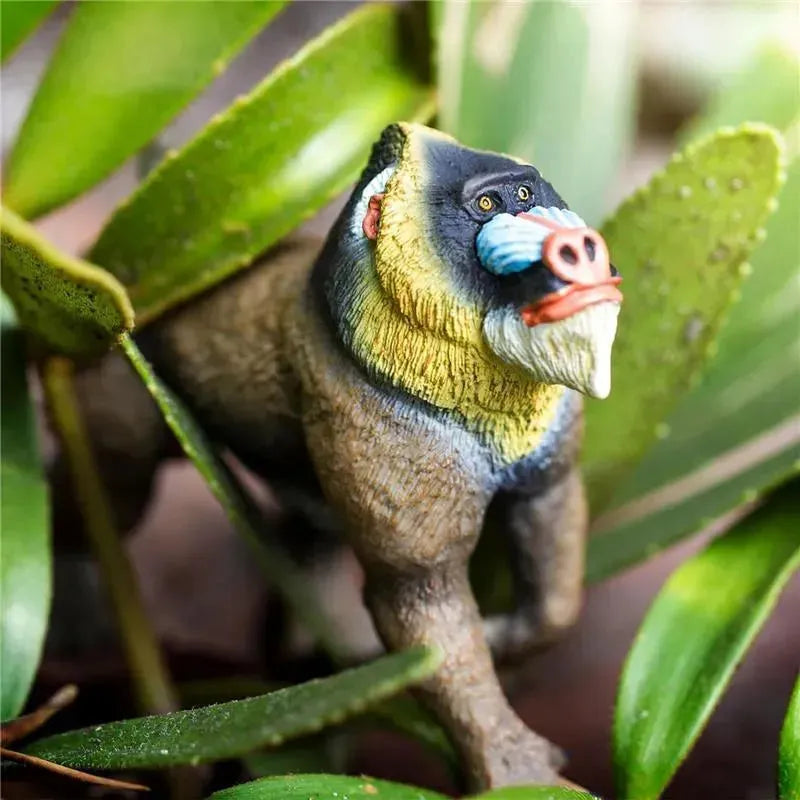 Mandrill figurine by Safari Ltd., featuring detailed colors and textures, set among green leaves.