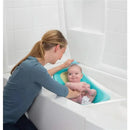Mother bathing a smiling baby in a blue and yellow infant bathing tub placed in a white bathtub.