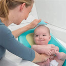 A parent gently brushes a smiling baby’s head with a cradle cap brush while the baby sits in a bath support.