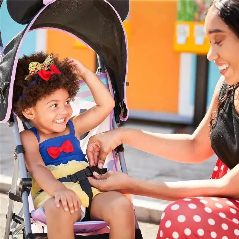 A young girl with curly hair smiles while sitting in a Disney Baby Mickey umbrella stroller, as a woman fastens her safety harness.