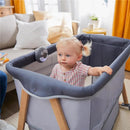 2-in-1 bassinet and play yard with a toddler inside, featuring soothing sounds, in a cozy living room setting.