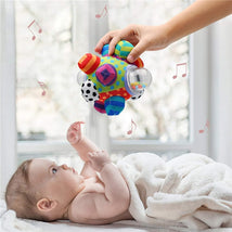 Baby reaching for a colorful Sassy Developmental Bumpy Ball held by an adult hand, with musical notes in the background.