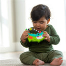 Toddler sits on a rug, holding a colorful Sassy Rock-A-Dile Musical Toy with buttons and beads, smiling and engaged.