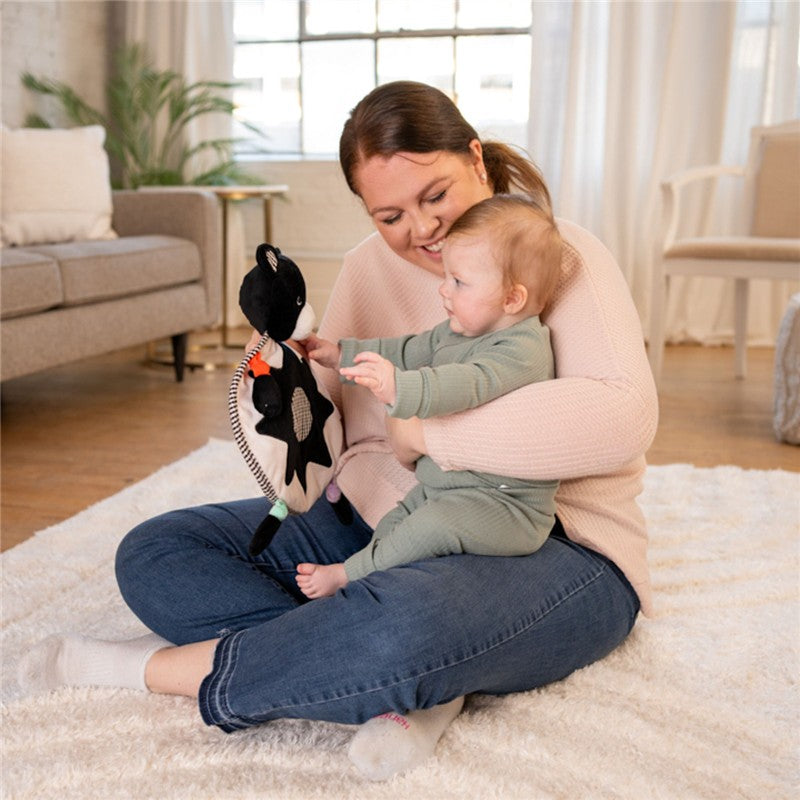 A woman sits on a rug, smiling and holding a Sassy Wimmer Bear Lovey while interacting with a happy baby.