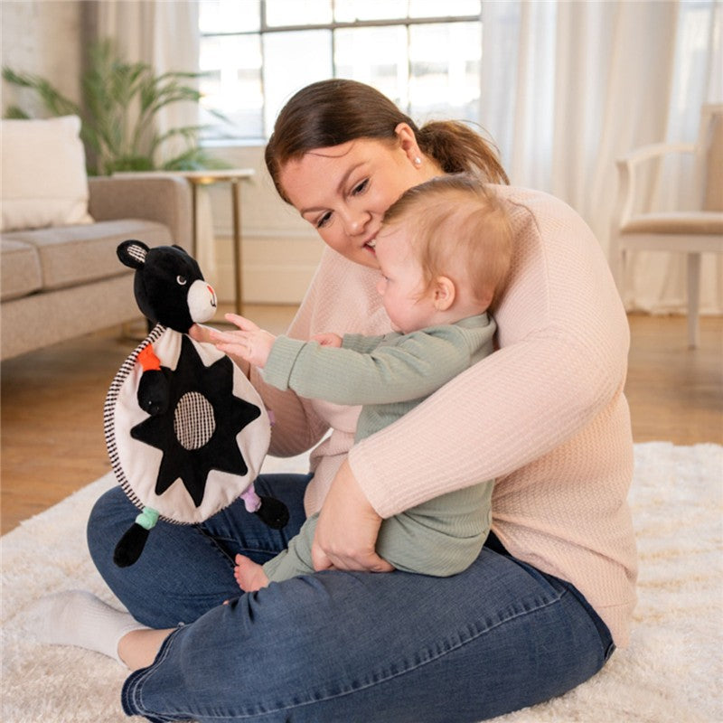 A mother and a toddler interact with a Sassy Wimmer Bear Lovey, featuring a black bear design and colorful accents.