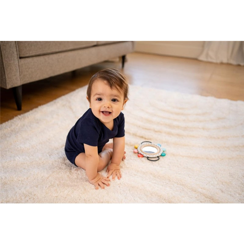 Smiling baby in a navy onesie, seated on a fluffy rug, with the Sassy Wimmer Grasp & See Mirror nearby.