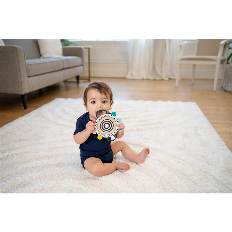 Infant sitting on a soft rug, holding a Sassy Wimmer Grasp & See Mirror toy, featuring colorful edges and black-and-white patterns.
