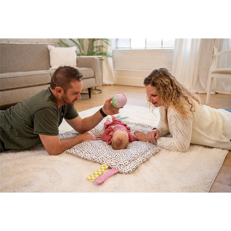 Parents interact playfully with a baby on a polka-dot mat, using a colorful toy to engage the child.