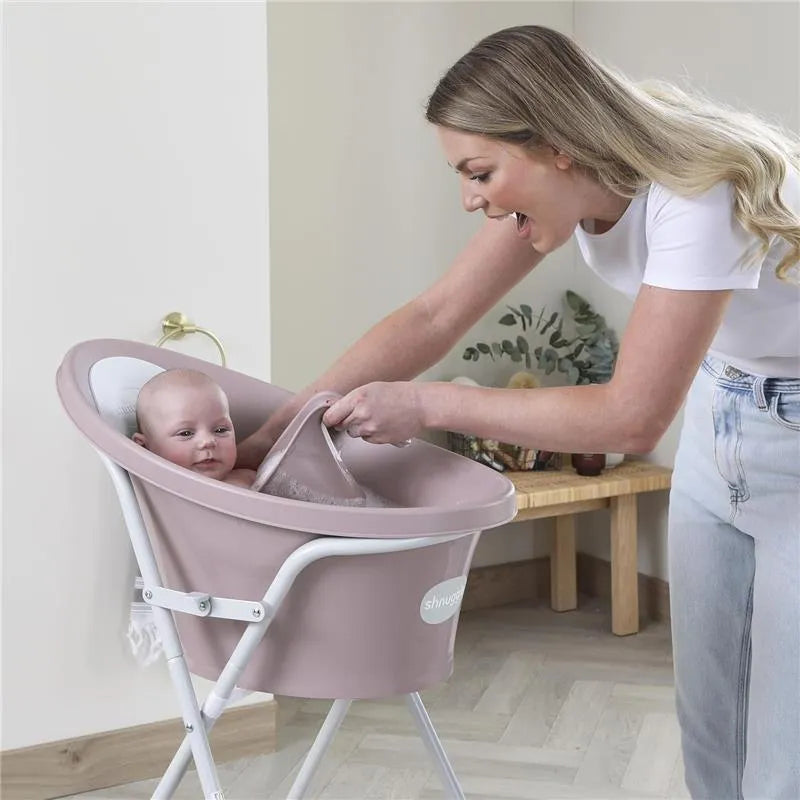 A woman using a Shnuggle bath rinse jug in a pink baby bath tub, gently rinsing a baby while smiling.