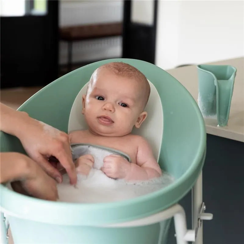 Baby bathing in a soft green Shnuggle bath, with a hand reaching to rinse using a eucalyptus bath rinse jug nearby.