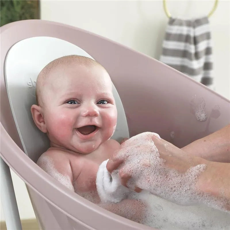 Newborn baby smiling while being bathed in the Shnuggle bath with bubbles and a towel in a cozy setting.