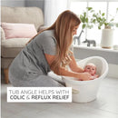 A caregiver bathes a newborn in the Shnuggle Newborn Baby Bath, highlighting its angled design for colic and reflux relief.