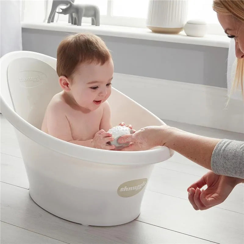 Newborn baby happily playing with a bath toy in a Shnuggle baby bath, while a caregiver assists.