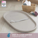 Squishy taupe baby changing diaper pad with a waterproof surface and safety strap, placed on a light wood floor.