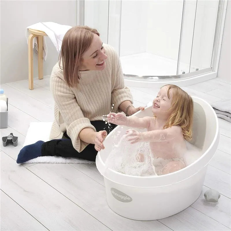 Toddler enjoys bathing in a Shnuggle white and grey tub while a smiling adult interacts playfully.