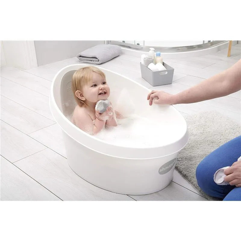 Toddler bathed in a white and grey Shnuggle tub, smiling and holding a toy, with a caregiver interacting nearby.