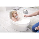 Toddler bathed in a white and grey Shnuggle tub, smiling and holding a toy, with a caregiver interacting nearby.
