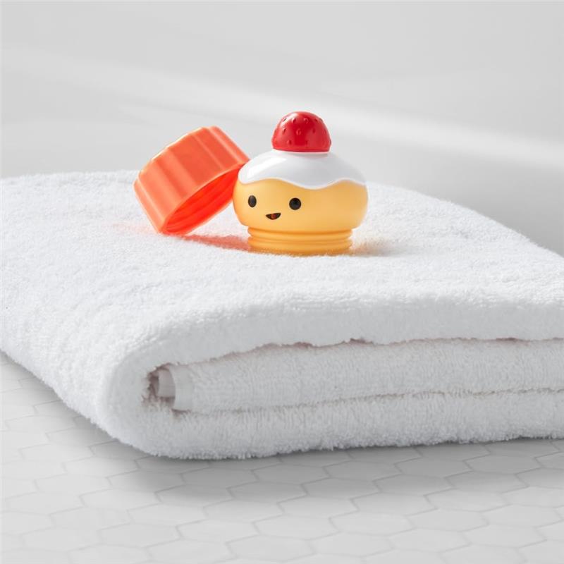 Bathtub toy resembling a cupcake with a removable lid, resting on a white towel, part of the Skip Hop Baby Bath Toy set.
