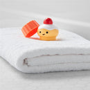 Bathtub toy resembling a cupcake with a removable lid, resting on a white towel, part of the Skip Hop Baby Bath Toy set.