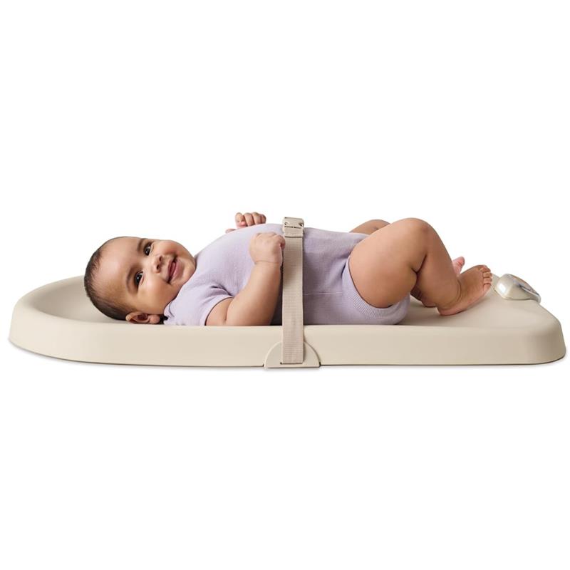 Baby lying on a light beige Skip Hop baby changing pad, smiling, secured by a strap, wearing a purple onesie.