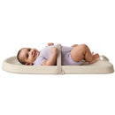 Baby lying on a light beige Skip Hop baby changing pad, smiling, secured by a strap, wearing a purple onesie.