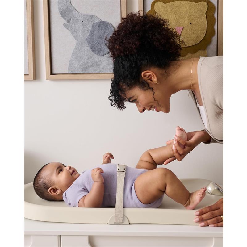 A mother smiling at her baby on a light-colored changing pad, engaged in playful interaction during a diaper change.