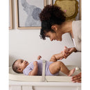 A mother smiling at her baby on a light-colored changing pad, engaged in playful interaction during a diaper change.