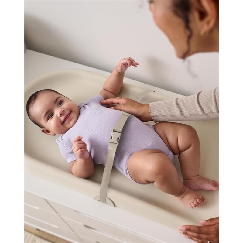 Baby lying on a Skip Hop changing pad, smiling, with a safety strap visible; caregiver touching the baby gently.