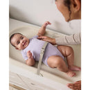 Baby lying on a Skip Hop changing pad, smiling, with a safety strap visible; caregiver touching the baby gently.