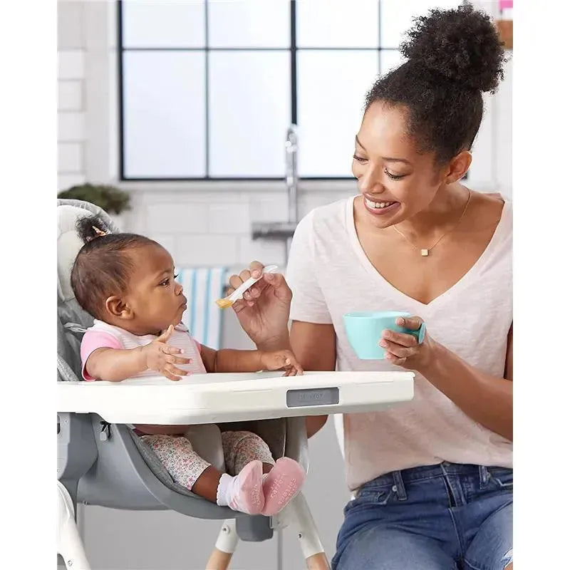 Mother feeding a baby in a high chair, using a spoon, with a teal feeding bowl in her hand.