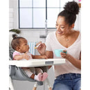 Mother feeding a baby in a high chair, using a spoon, with a teal feeding bowl in her hand.